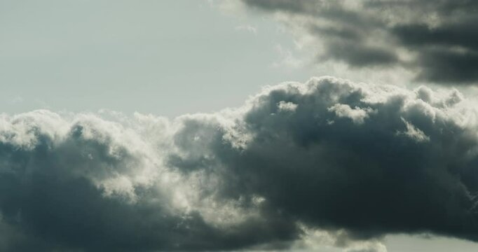 Timelapse of close-up view of contrasting dark gray fluffy cumulus clouds with bright white edges rolling across the pale colored sky