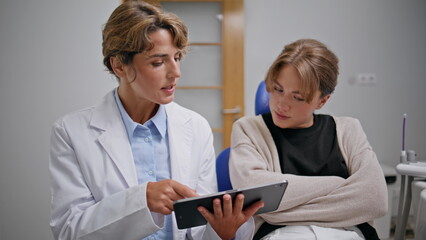 Stomatologist explaining medical treatment on tablet to woman in clinic closeup.