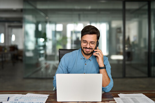 Smiling Hispanic businessman making call on mobile cellphone, consulting client having work conversation. Busy happy young Latin business man talking on phone using laptop computer in office.
