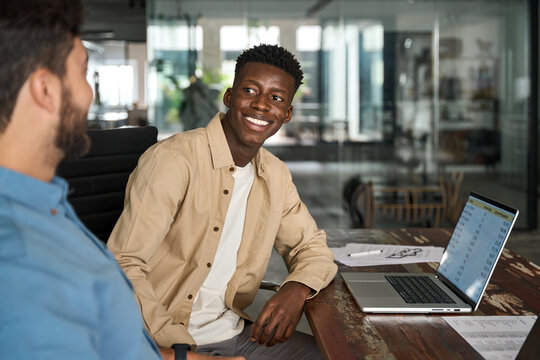 Happy African male employee having conversation with Latin colleague at work. Smiling friendly diverse workers team talking working together using laptop computer sharing ideas at workplace in office.