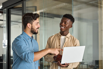 Business team of two busy happy professional business men employees working together standing in corporate office having conversation sharing ideas using laptop computer technology.