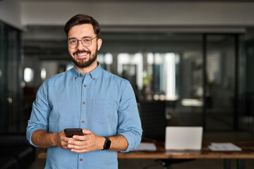 Happy young Latin business man holding phone standing at work in office. Smiling professional...