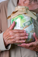 An elderly persons hands holding a glass jar filled with various bills, wrapped in a paper bag