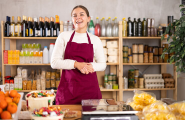 Fototapeta premium Young female seller in apron standing at counter in grocery store