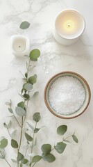A bowl filled with sea salt lies next to fresh eucalyptus leaves and a softly glowing white candle all beautifully arranged on a white marble surface to enhance a calming spa ambiance