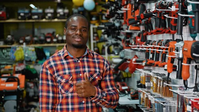African-American man in a plaid shirt gives a thumbs-up while standing in a hardware store aisle filled with drills and power tools