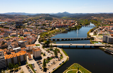 Picturesque aerial view of Mirandela city with residential houses with brownish tiled roofs, bridges crossing Tua river and water fountain on spring day, Braganca, Portugal