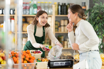 Grocery store selling area. Female employee sells candies to woman, puts them in bag, puts them on scale and weighs sweets. Brunette European girl buys candies by weight.