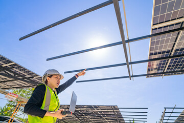 A technician in a safety helmet and reflective vest inspects a solar panel structure using a...