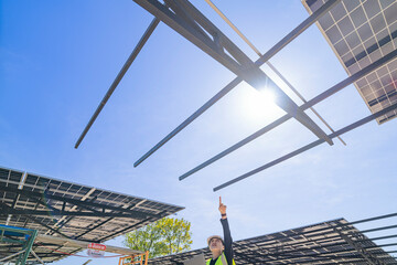 A technician in a safety helmet and reflective vest inspects a solar panel structure using a laptop. The solar panels are mounted on a steel carport framework under a clear blue sky.