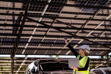 Fototapeta premium A female technician in a hard hat and reflective vest conducts a solar panel inspection with a laptop and a handheld device under a solar carport structure. The panels and sunlight frame the scene.