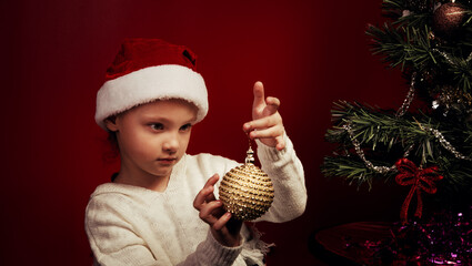 Smiling cute with thinking magic eyes kid girl holding in hands Christmas tree golden ball near the holiday fur green tree in red santa claus hat. Happy expression winter holadays. Closeup
