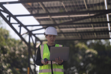 A female technician wearing a safety helmet and reflective vest uses a laptop while inspecting a solar carport installation. The scene features solar panels and parked cars under a clear sky.