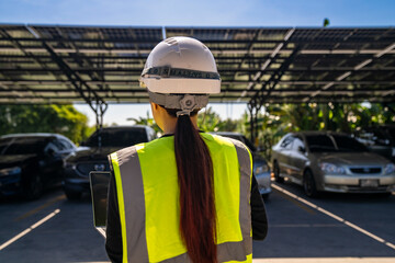 Female engineer in safety gear and helmet inspecting a solar-powered carport. She observes parked vehicles shaded by photovoltaic panels, promoting clean energy technology and sustainability.