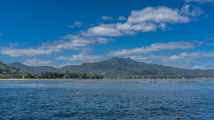 Mountains against the blue sky, clouds. On the coast there is green vegetation and city buildings. Yachts, boats are moored. Silhouettes of fishermen in shallow water. The ocean is in the foreground