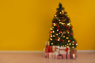 Blurred view of Christmas tree with glowing lights, ornaments and gift boxes near yellow wall