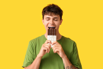Handsome young man eating sweet chocolate bar on yellow background