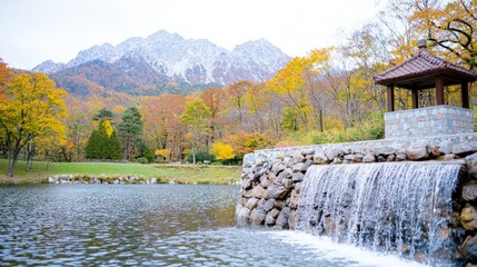 Naklejka premium Beautiful waterfall flowing from a stone wall into a calm pond surrounded by colorful autumn trees and majestic mountains.