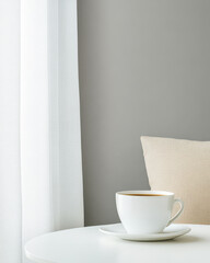 Coffee cup on a white table near a window with soft curtains and a beige cushion