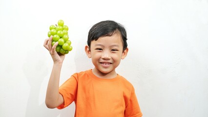 A cheerful young boy in an orange shirt holding a bunch of fresh green grapes, smiling brightly against a clean white background. The image radiates happiness, health, and freshness of natural fruit.