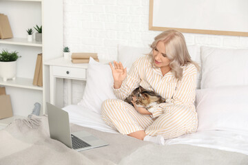 Beautiful mature woman in pajamas with cute cat and laptop video chatting on bed at home