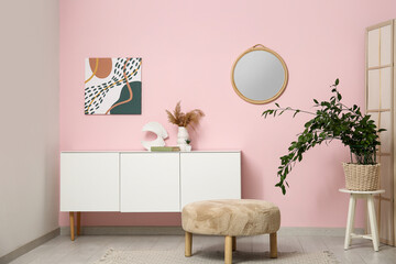 Chest of drawers, pouf, houseplant, picture and mirror on pink wall in room