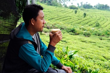 A man enjoys a hot drink while overlooking a lush green tea plantation. A serene moment in nature,...