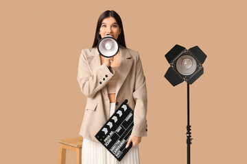 Young shocked female film director with movie clapper, megaphone and studio lighting on beige...