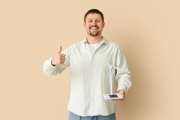 Young man with wind turbine model showing thumb-up on beige background. Green energy concept