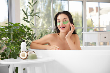 Beautiful young happy woman with under-eye patches, cosmetic products and coconut sitting in bathtub at home