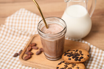 Glass of sweet chocolate milk with cocoa beans and tasty cookies on wooden background