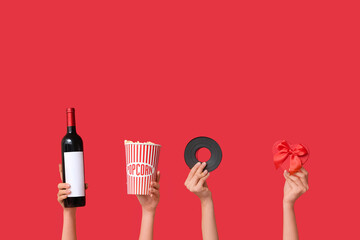 Female hands with bottle of wine, bucket of popcorn, film reel and gift box in shape of heart on red background. Valentine's Day celebration