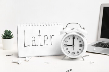 Notebook with word LATER, alarm clock, earphones and laptop on light wooden table