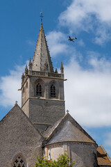 Obraz premium Church steeple with a plane flying in the sky. The plane is flying low and is in the background. Saint Come du Mont, normandy, France