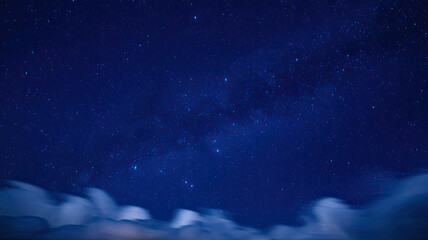 Fototapeta premium Vast Star-Studded Night Sky with Deep Blue Hue, Bright and Luminous Stars Scattered Across, Soft Fluffy Clouds at the Bottom Adding Depth and Contrast, Creating a Serene and Enchanting Nighttime Atmos