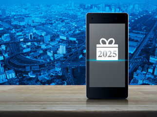 Gift box happy new year 2025 flat icon on modern smart mobile phone screen on wooden table over...