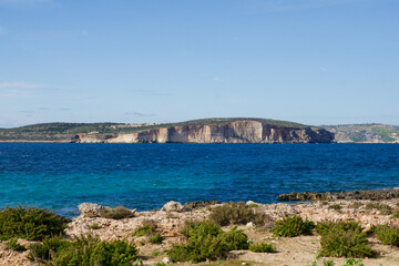 Malta, January 1, 2020: Scenic Coastal View with Green Bushes, Turquoise Mediterranean Sea, and Dramatic Cliffs of a Nearby Island