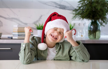 Portrait of happy boy in Santa Claus hat in the kitchen with red Christmas decor, smiling in anticipation of the holiday. New Year.