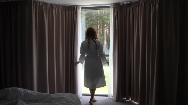 Slow motion of woman in robe opens curtains in room, revealing forest view outside. Wake up at the morning, weekend vacation on nature. Designed luxury home interior. self-care, new day starting