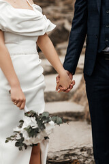 two figures holding hands. The woman is wearing a white dress with drapery on the shoulders and holding a bouquet of white flowers with green leaves. The man is wearing a dark suit.