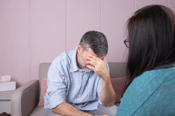 Fototapeta premium A man in a therapy session hides his face with one hand, while his psychotherapist, with his back turned, accompanies him in a quiet environment. Concept of therapy and mental health
