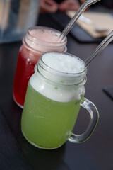 Deux pichets de boissons fraiches colorées avec de la mousse épaisse et des pailles en verre. Cocktails posés sur une table sombre dans un restaurant.
