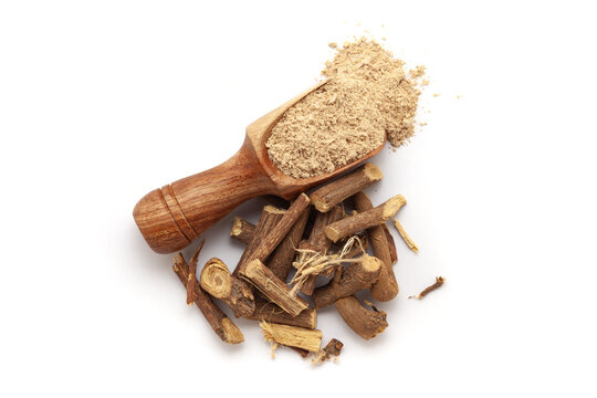 A wooden scoop filled with organic Liquorice or Mulethi (Glycyrrhiza glabra) powder, placed beside a heap of liquorice roots, isolated on a white background.