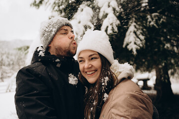 A young man wearing a gray hat covered with snow blows snow off the white hat of his smiling wife. In the background are large fir trees in the snow. Happy winter journey.