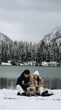 A happy family of three sits in the snow by the lake, all dressed in warm winter clothes. In the background, majestic mountains are covered with snow.