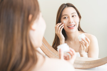 Facial beauty skin care, smile of pretty asian young woman in bathrobe looking at mirror, hand applying moisturizer lotion on her face, holding jar of skin cream before makeup cosmetic routine at home