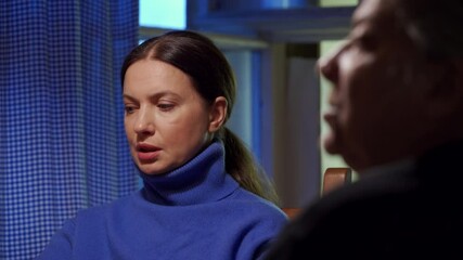 Adult 44 year old woman having lively conversation with elderly woman sitting at table in living room. Woman with an upset, tense face listens and talks.