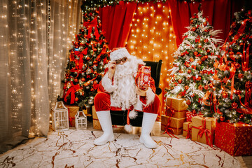 Santa Claus holding a wrapped gift and adjusting his sunglasses, surrounded by decorated Christmas trees, glowing lights, and presents.