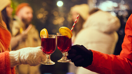 Two glasses of mulled wine with orange slices and striped straws clinking together, celebrating cozy winter evening under festive lights. Friends gathering together. Winter holidays, Christmas concept