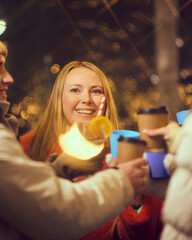 Happy young woman with long red hair holding glass of mulled wine, surrounded by friends and illuminated by warm, festive lights at a holiday fair. Concept of winter holidays, Christmas, traditions